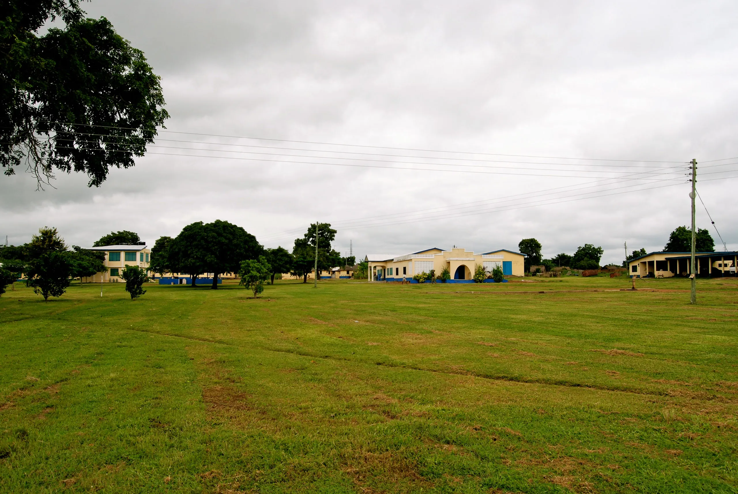 A wide-angle view of the Carpenter project site in Ghana, showing single-story buildings with white walls and blue trim set against a expansive green lawn under an overcast sky.