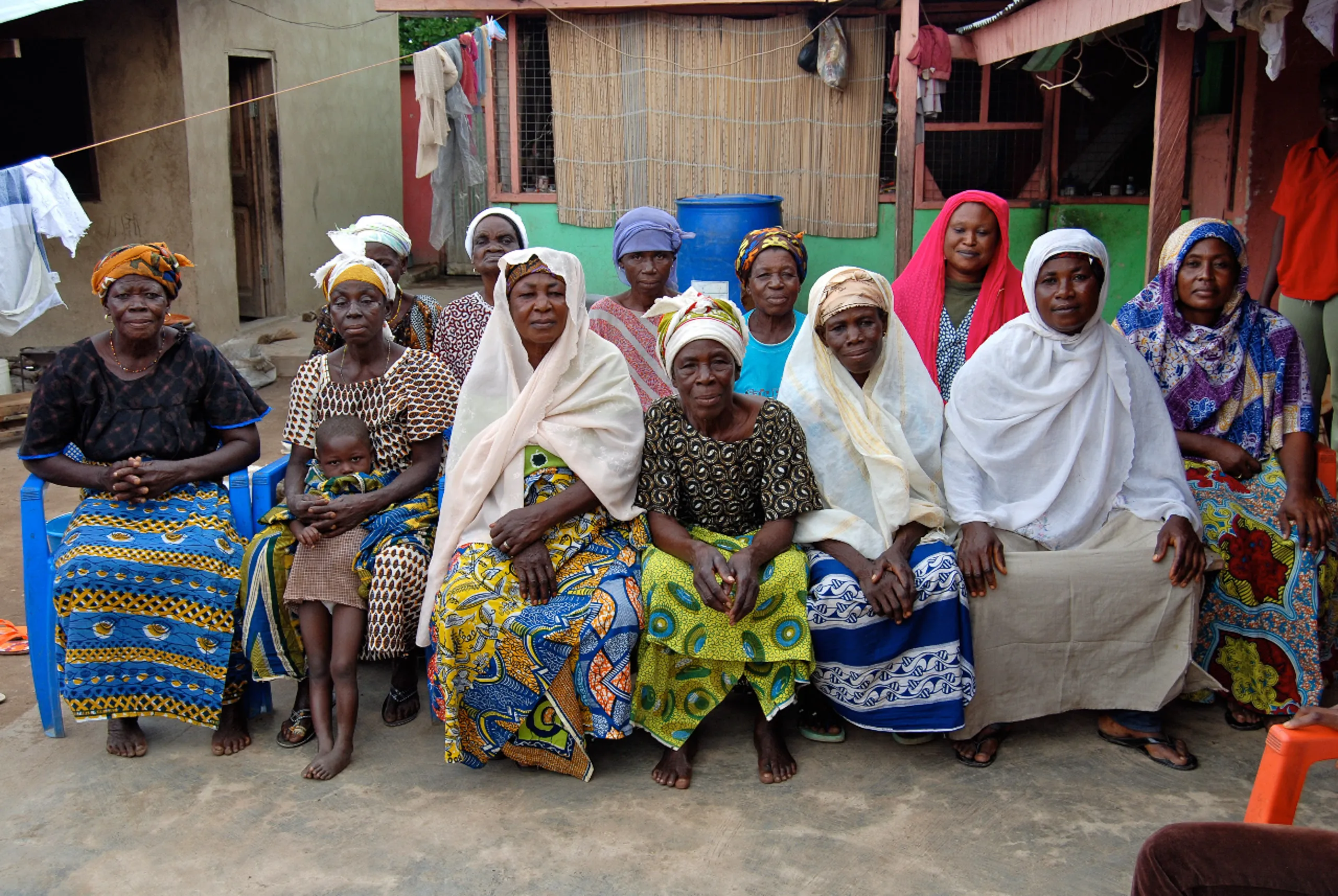 A group portrait of women sitting outdoors together, wearing colorful patterned skirts, headwraps, and traditional garments, with one woman holding a young child.