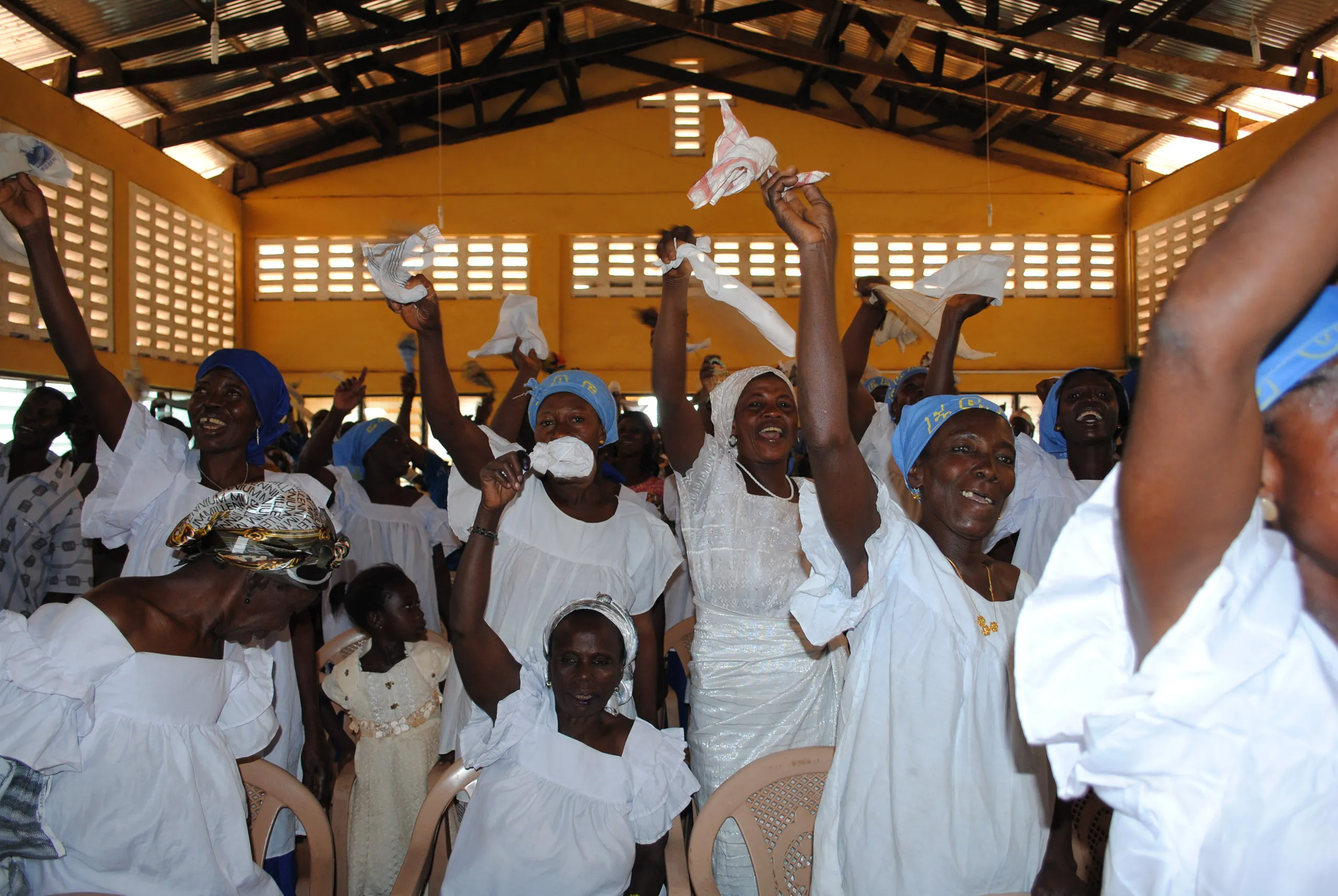 A joyful church congregation inside a building with a yellow wall and open-slat windows, with women in white clothing raising their hands and waving white cloths in celebration.