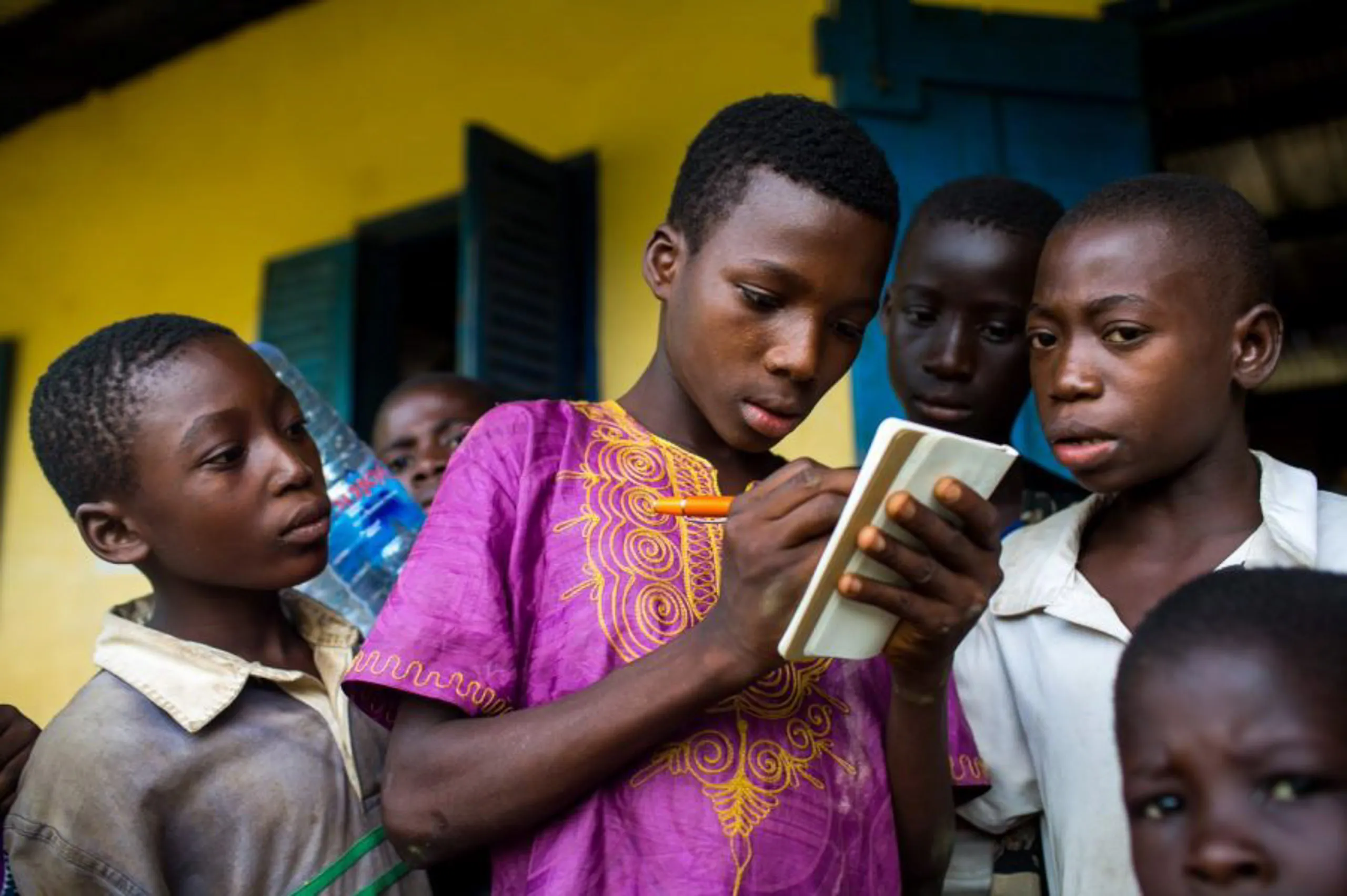 A young student in a purple patterned tunic uses a pen and small notebook while other children look on curiously.