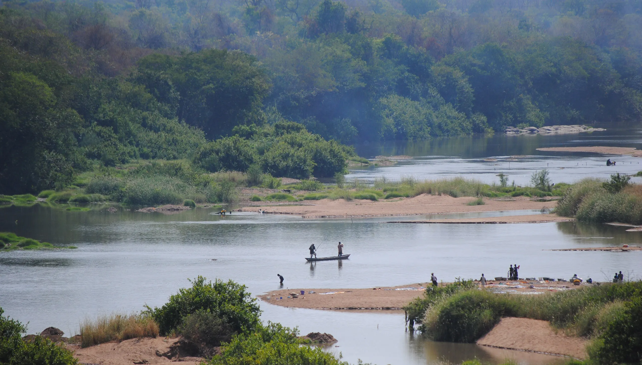 A scenic, elevated view of the Black Volta River winding through lush green forests with small groups of people near the water's edge, including a small boat with two fishermen.