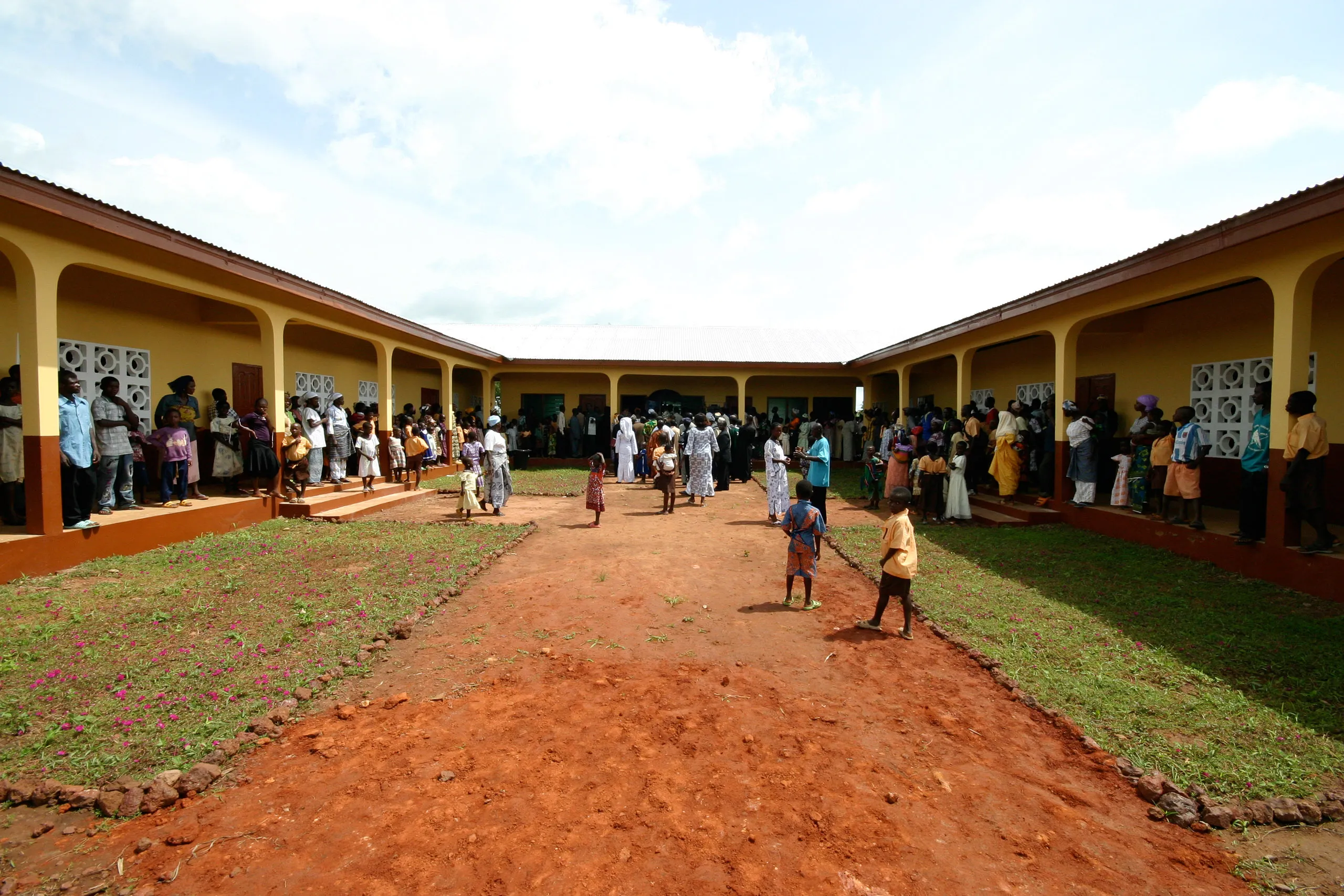Courtyard view of a U-shaped school building with covered archways, surrounded by people walking on a red dirt and grass courtyard.