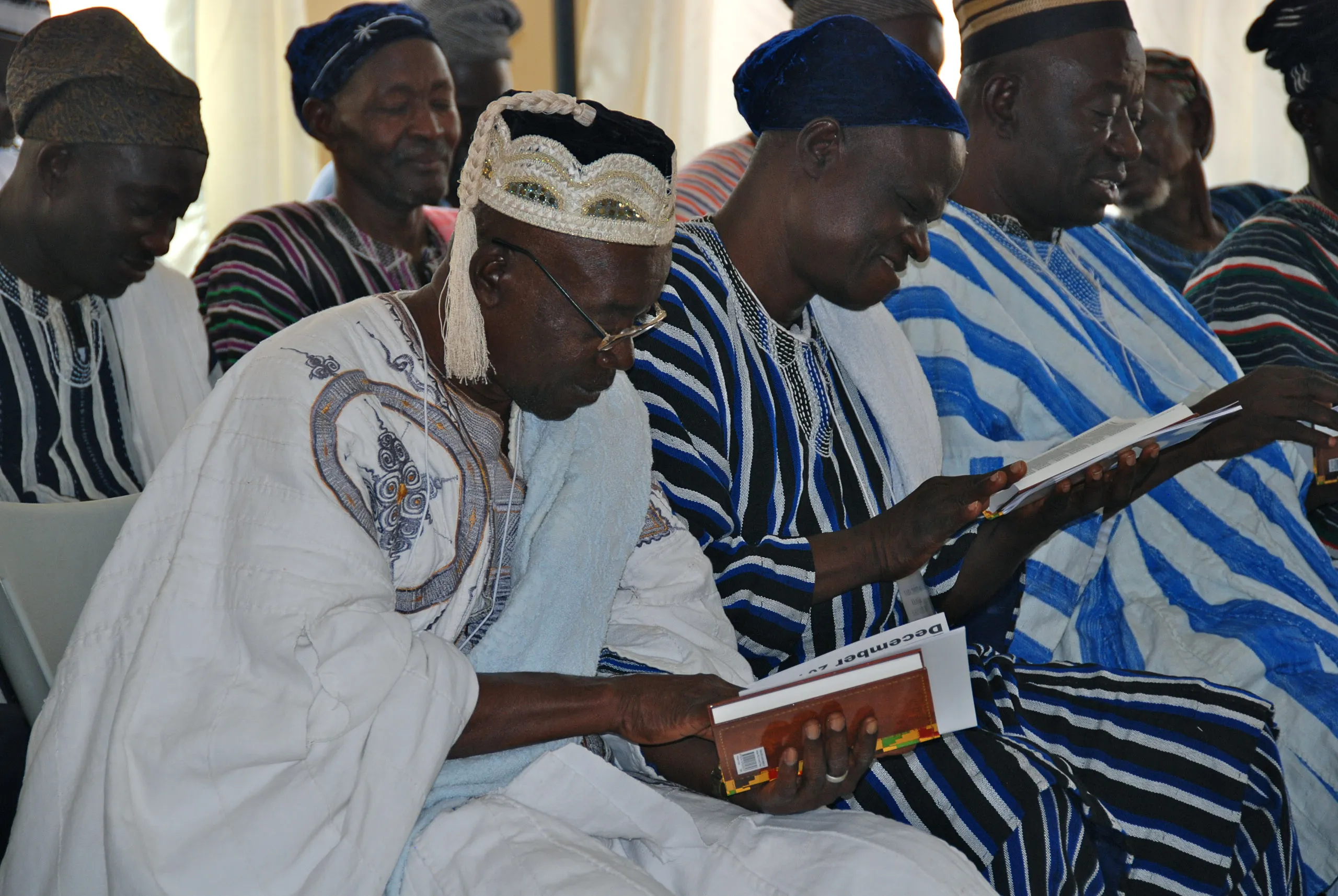 Several Ghanaian chiefs wearing traditional smocks and patterned caps attentively read from materials at a meeting.