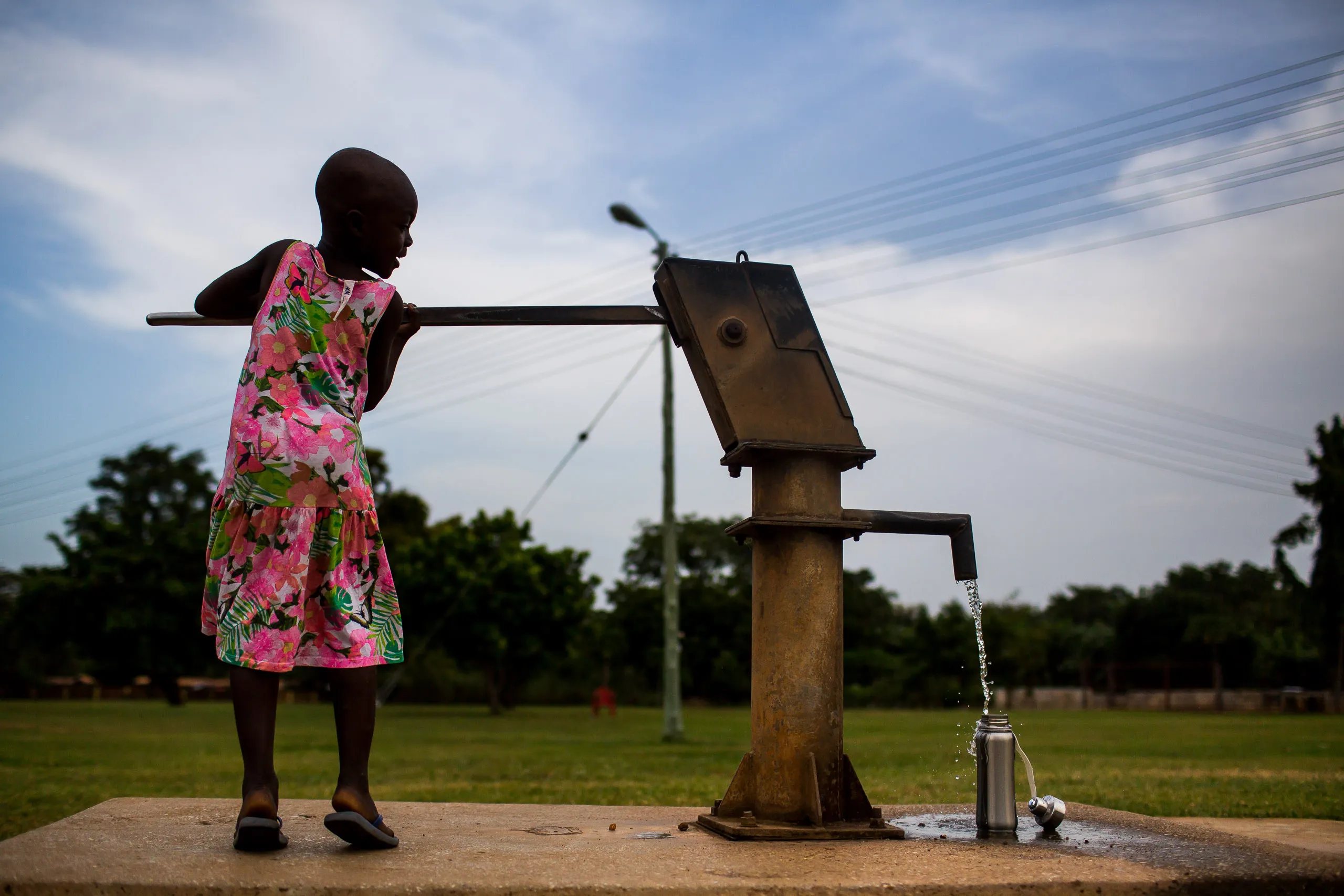 A young girl in a pink floral dress operates a hand pump at a water well, filling a metal bottle placed on the concrete base.