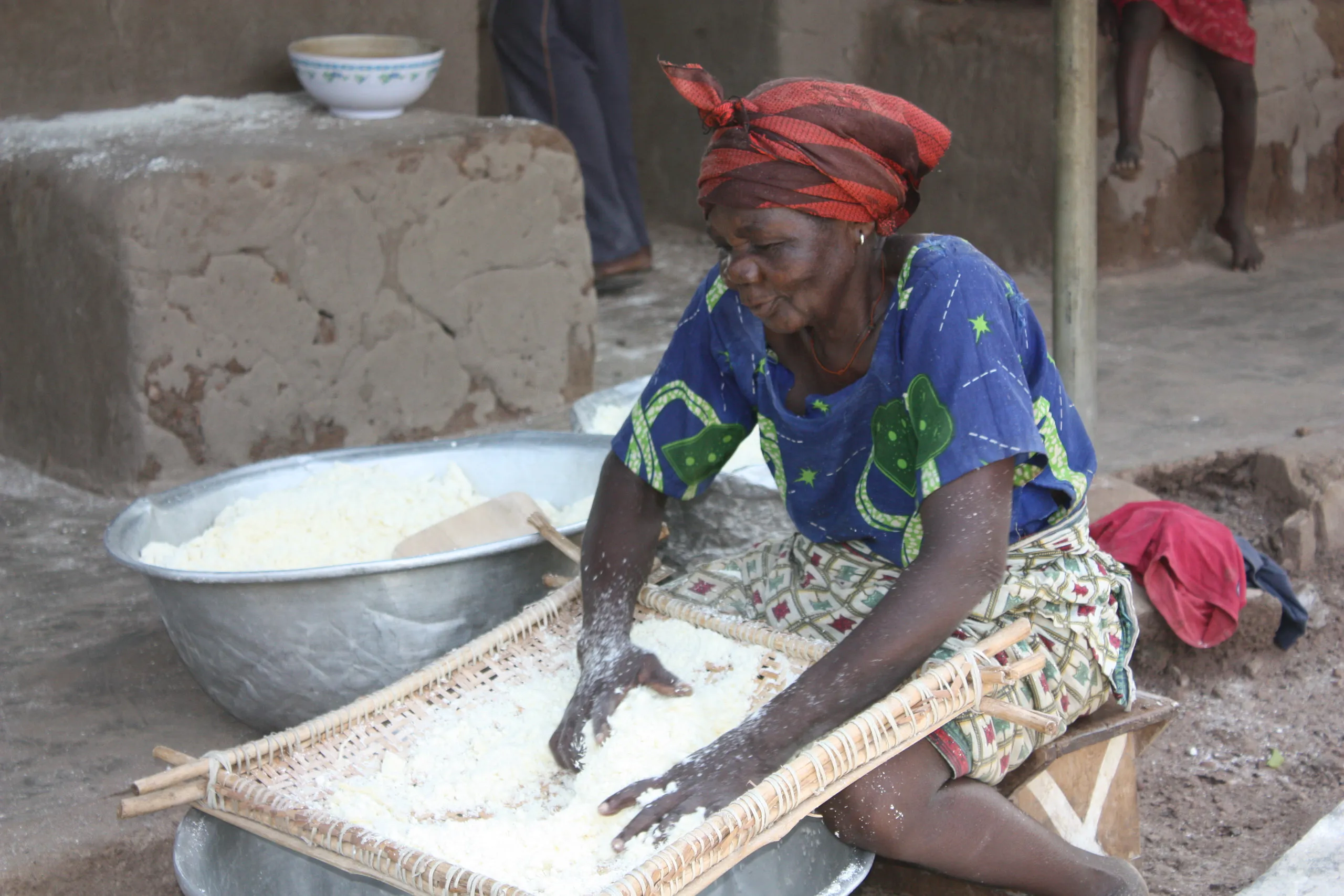 An elderly woman wearing a blue patterned top and a red headwrap sits on the ground and manually sifts grated cassava through a woven tray.
